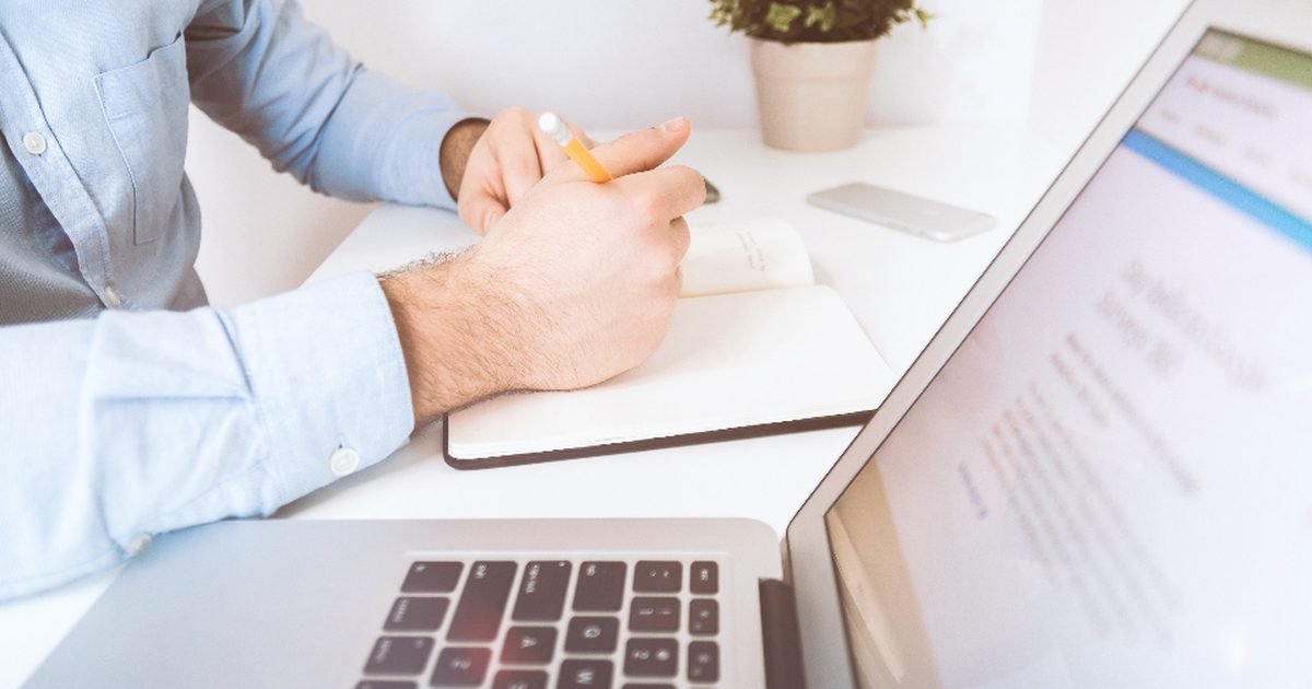 Person writing meeting notes beside a laptop during a work session