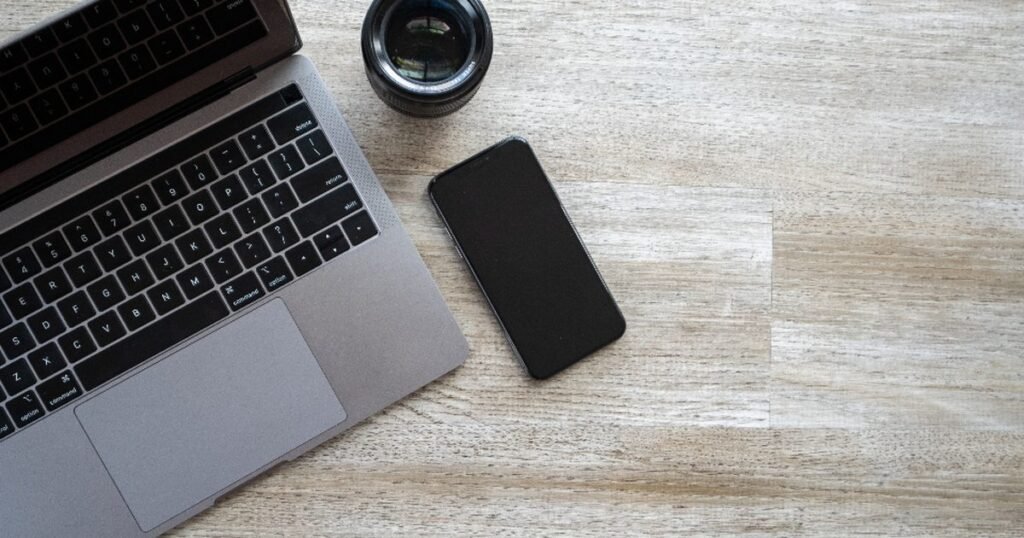Smartphone beside a laptop and camera lens on a desk for a phone storage cleanup guide